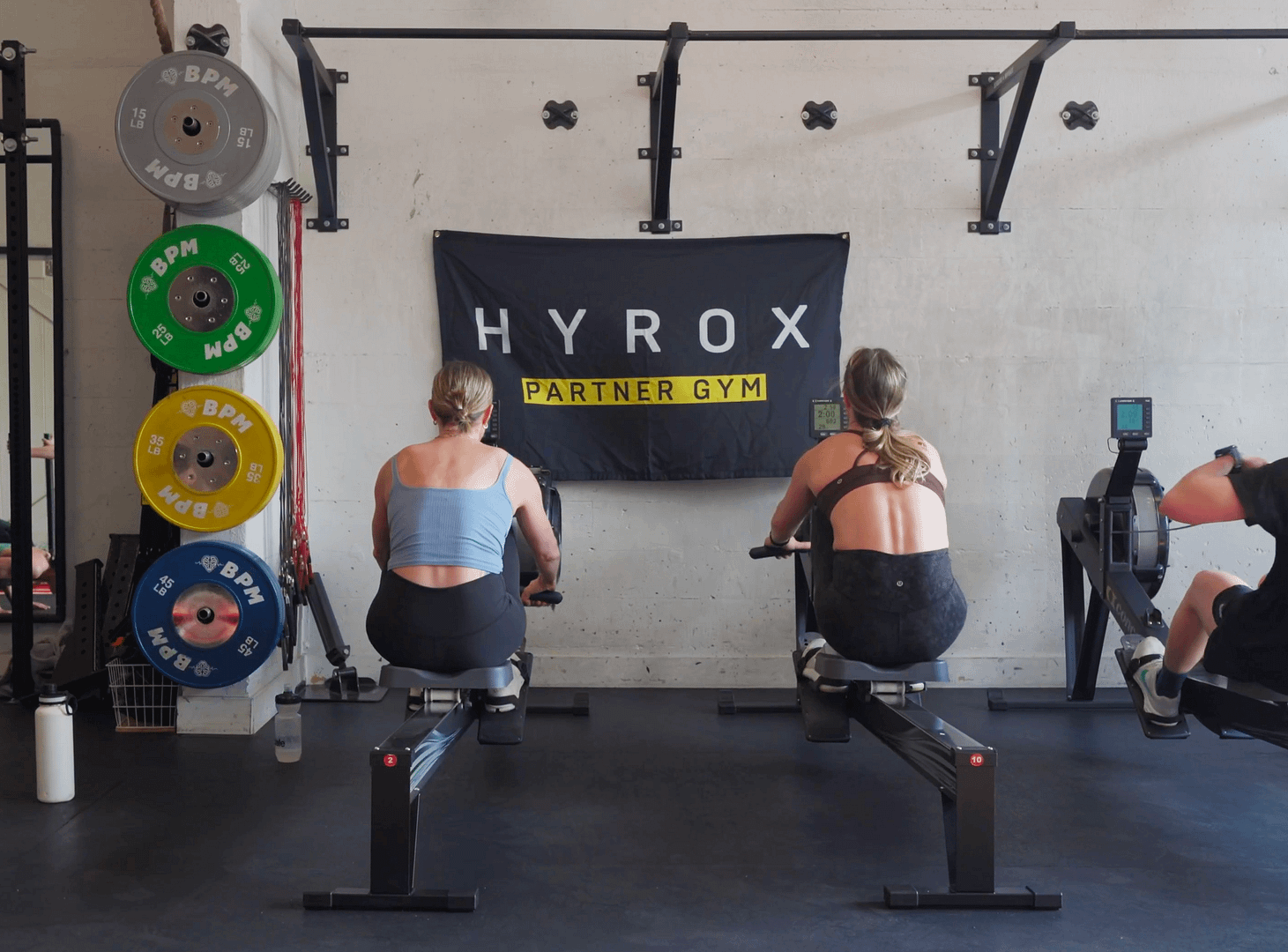 Two girls on Concept 2 Row Ergs with a HYROX flag in the background at BPM Fitness Centre