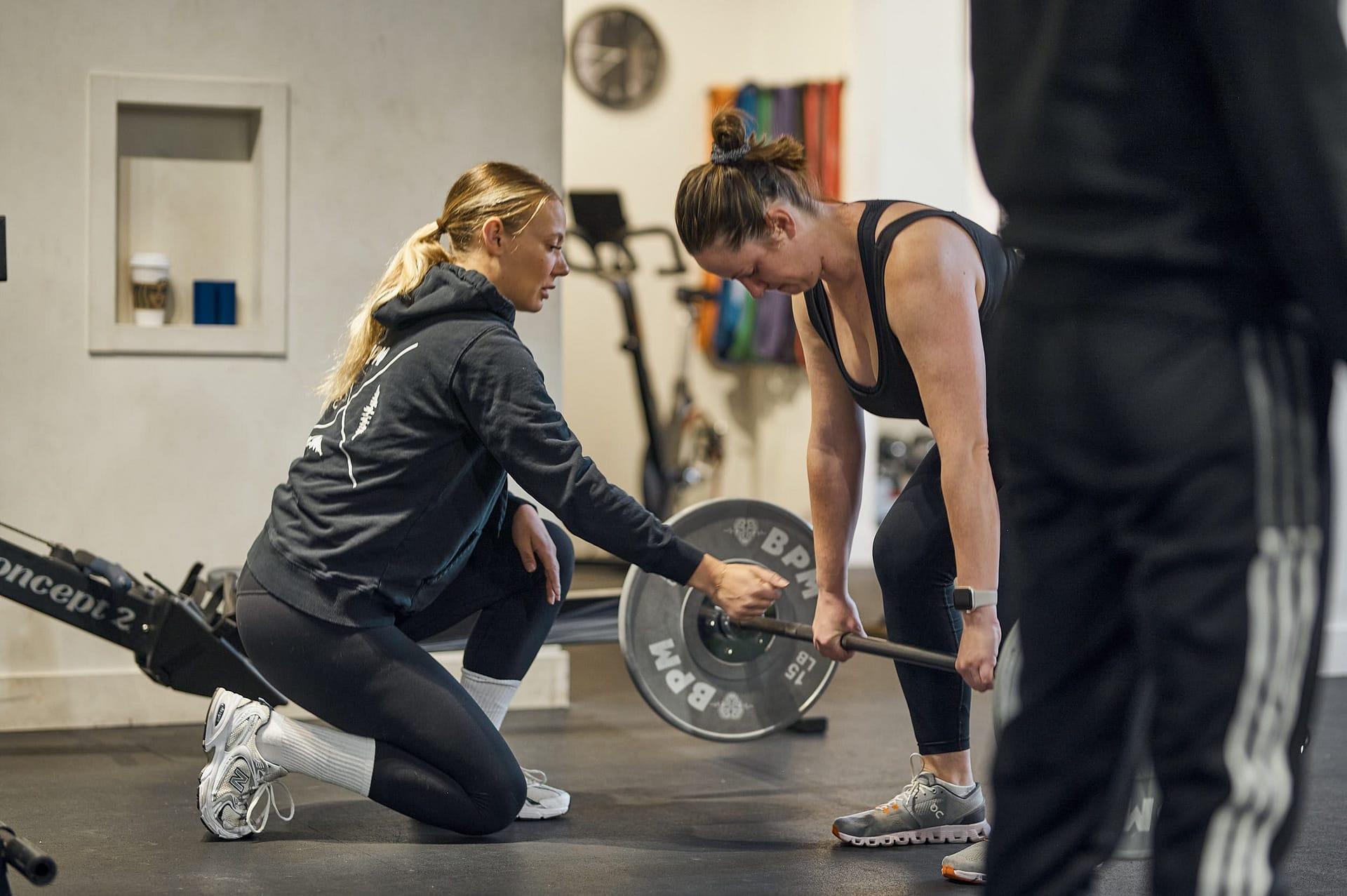 Two women engaged in a personal training session, focusing on weightlifting with a barbell in a gym setting.