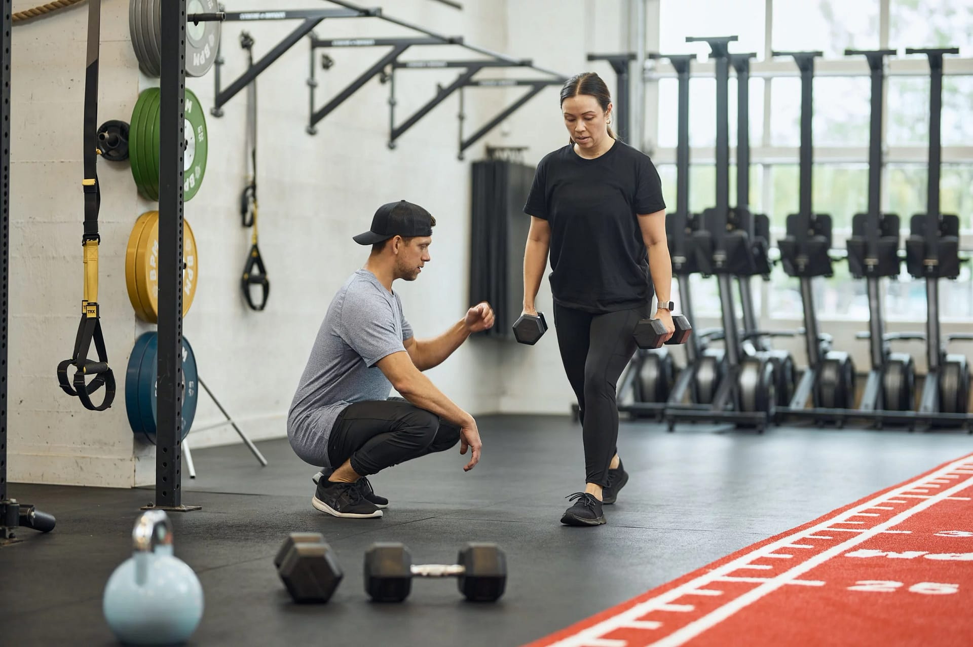 A personal trainer assists a woman with dumbbell exercises in a gym setting, showcasing fitness training and strength building.